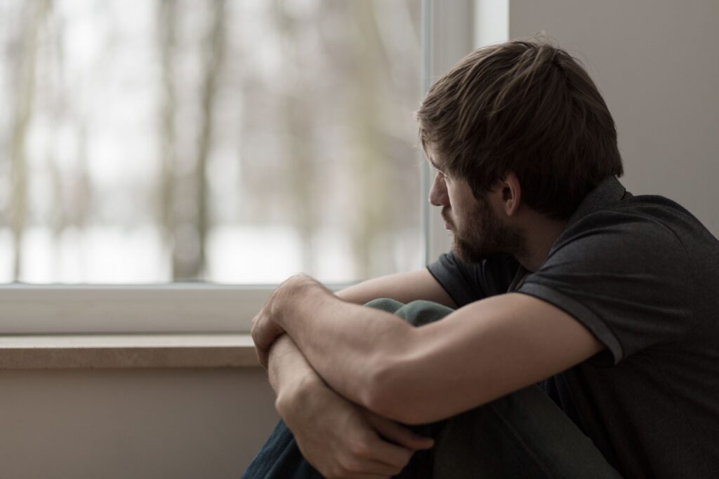 Depressed man with arms around knees gazing out window
